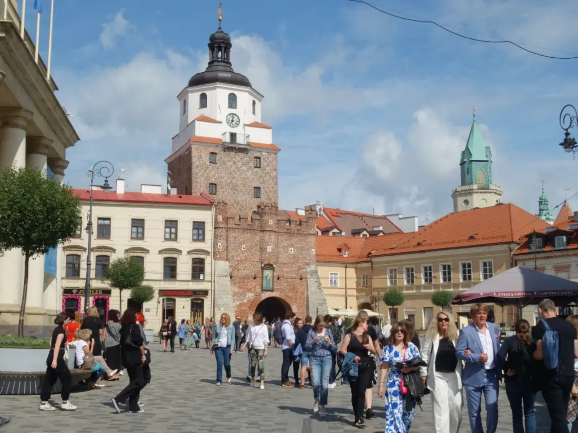 Ludzie spacerują po brukowanym placu litewskim, podziwiając zabytkową wieżę z zegarem i zieloną iglicę w tle.