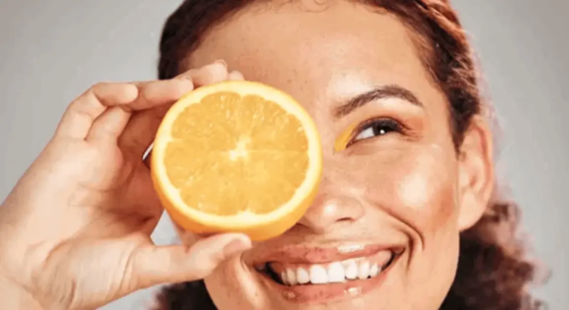 Strahlendes Lächeln mit einer Orangenscheibe vor dem Auge, die Frische und Vitamin C für die Hautpflege symbolisiert.