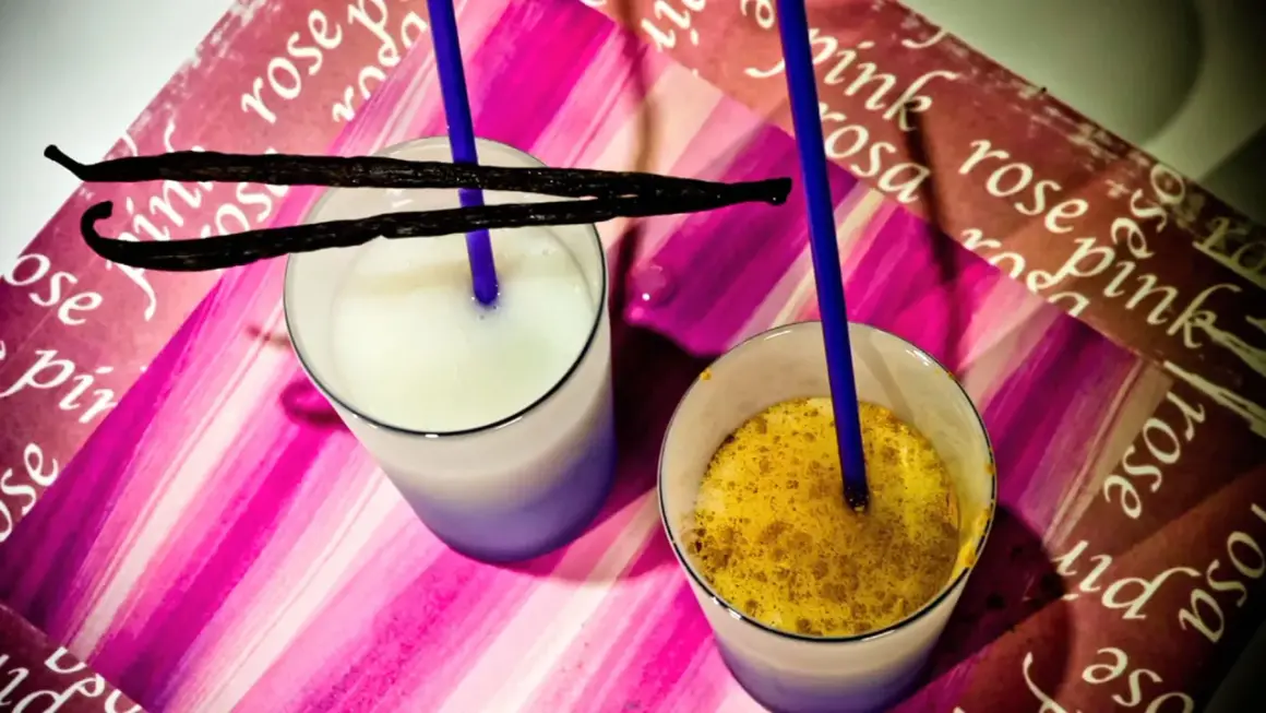 Due bicchieri con latte di riso, uno bianco e uno con spezie dorate, adornati da baccelli di vaniglia e cannucce blu su sfondo rosa.