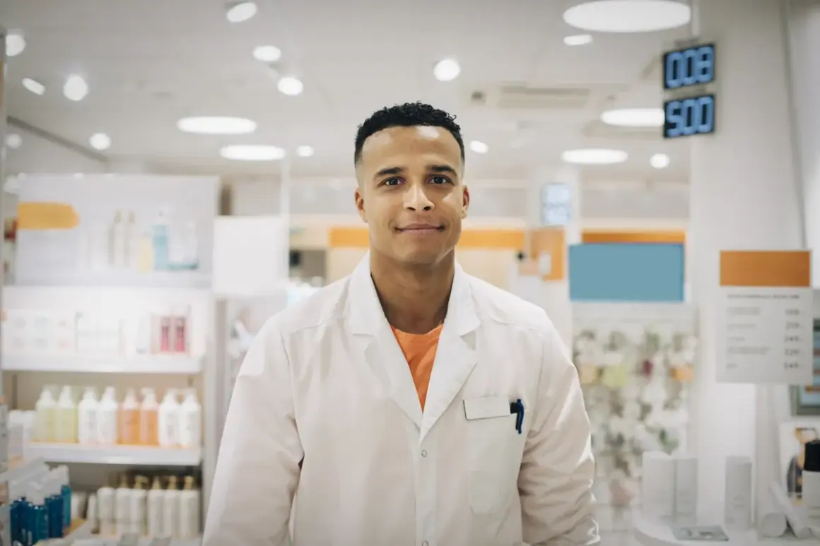 Ung man i vit rock, redo för apotekstekniker utbildning. Bakom honom syns hyllor med produkter i ett apotek.