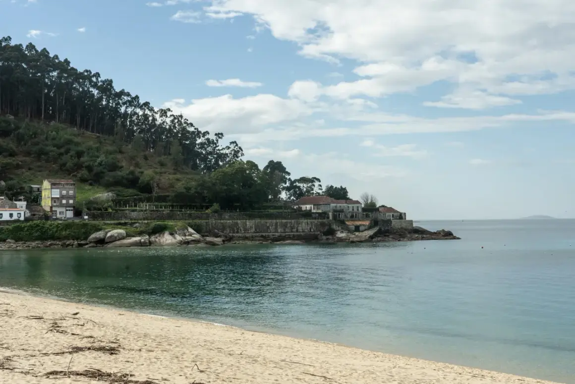 Arena dorada y mar turquesa en la playa de Loira, con casas blancas y una colina boscosa al fondo.