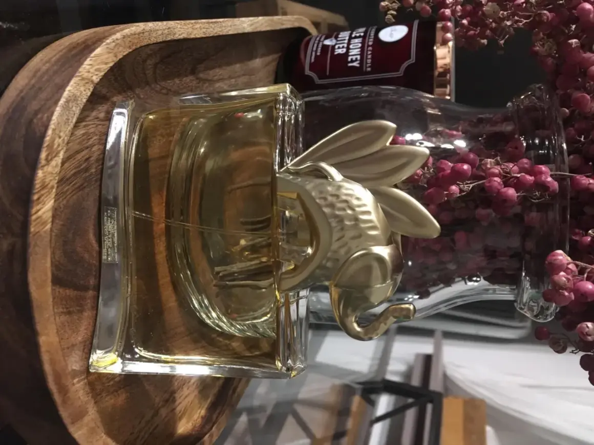 Złoty słoń na flakonie Kenzo Jungle 100ml, otoczony drewnianą misą i różowymi owocami.