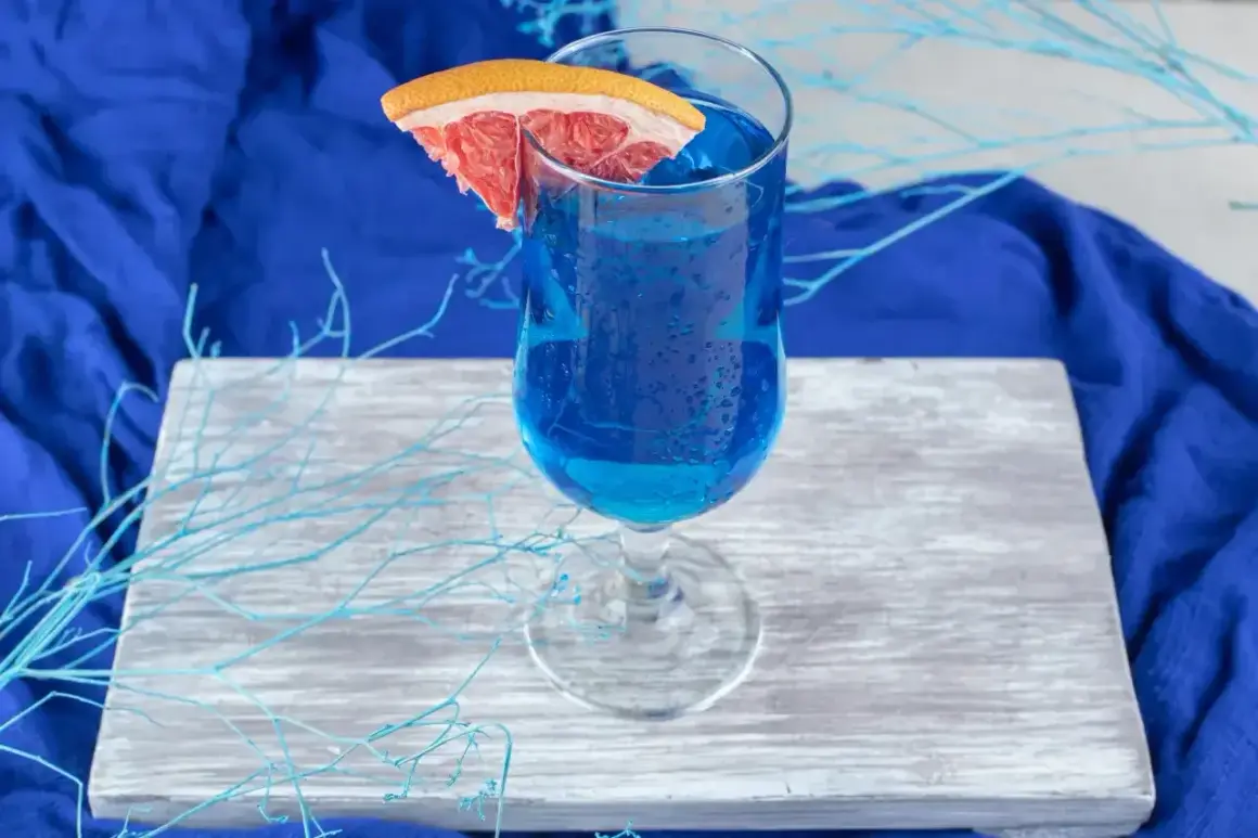 Niebieski drink, jak orzeźwiająca blue lagoon, z plasterkiem grejpfruta, na drewnianej desce, otoczony błękitnymi gałązkami.