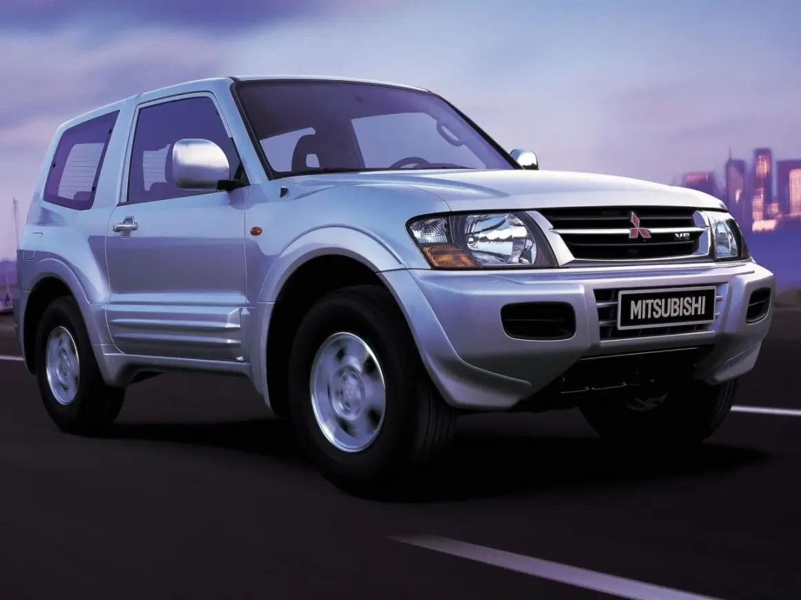 Srebrny Mitsubishi Shogun 3-drzwiowy na drodze o zmierzchu, z miejskim krajobrazem w tle.