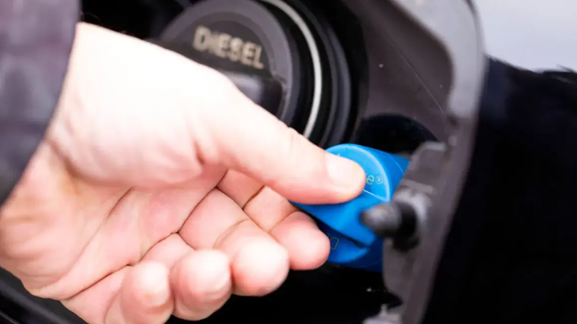 Hand schraubt blauen AdBlue-Deckel ab. Diesel-Tankdeckel daneben.