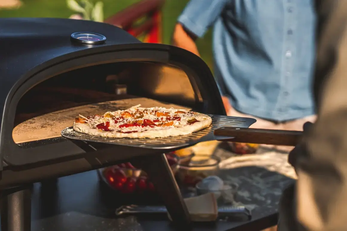En pizza på muurikka är på väg in i en utomhusugn. Solen skiner och en person i blå skjorta står i bakgrunden.