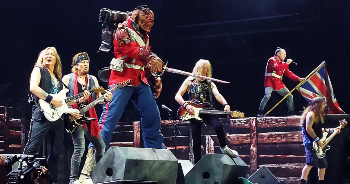 Zespół Iron Maiden na scenie, w tym Eddie w czerwonym mundurze, grający na gitarach i z flagą Wielkiej Brytanii.