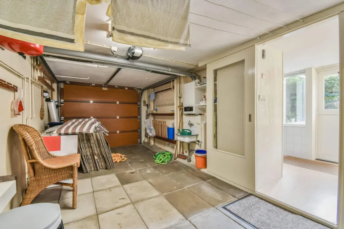 Un garage en cours de transformation en habitation. On y voit une porte de garage, un évier, une chaise en rotin et des éléments de rangement.