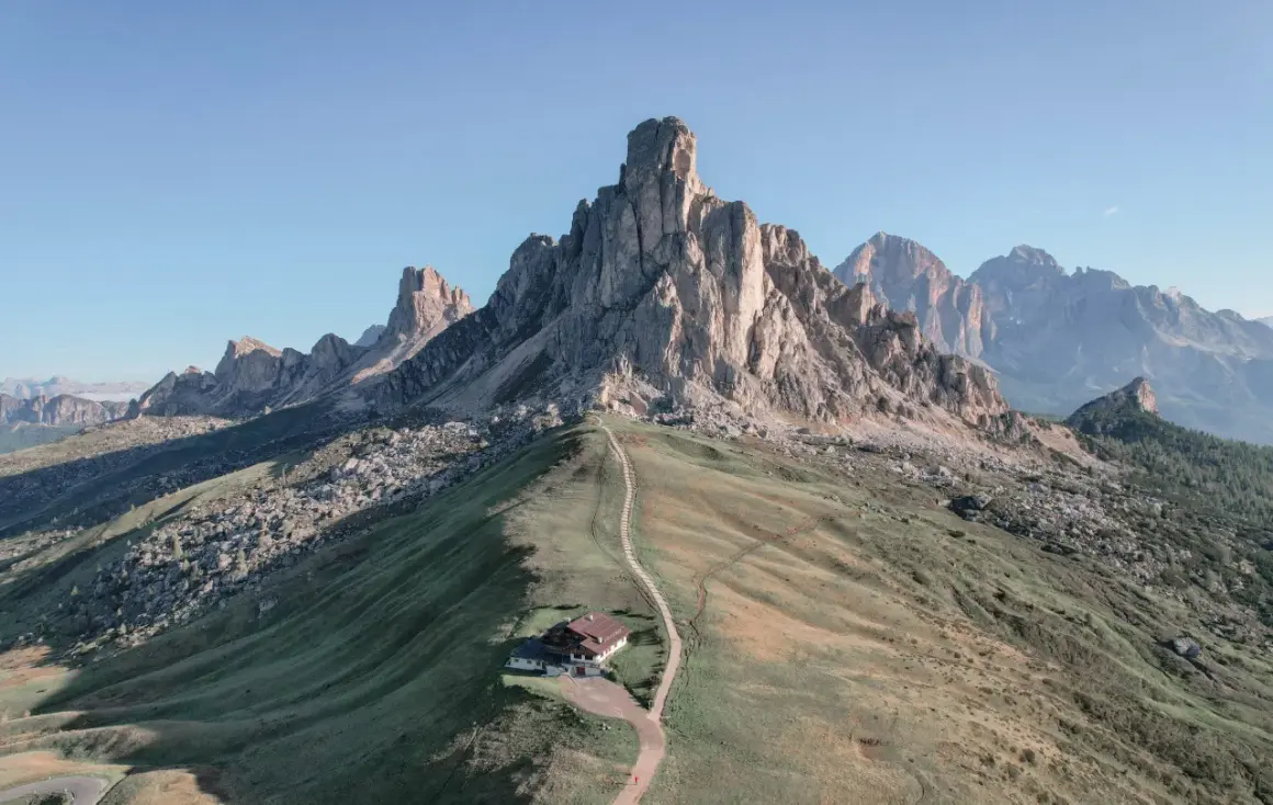 Un refugio alpino solitario en la ladera de una montaña, con un sendero que serpentea hacia él. El paisaje evoca la belleza de **Milano Cortina**, con picos imponentes y cielos despejados.