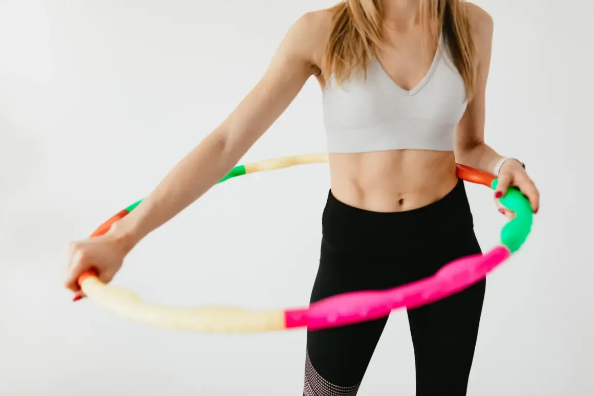 Kobieta w sportowym topie i czarnych legginsach kręci kolorową hula hop. Widać jej wyrzeźbiony brzuch, co sugeruje pozytywne kręcenie hula hop efekty.