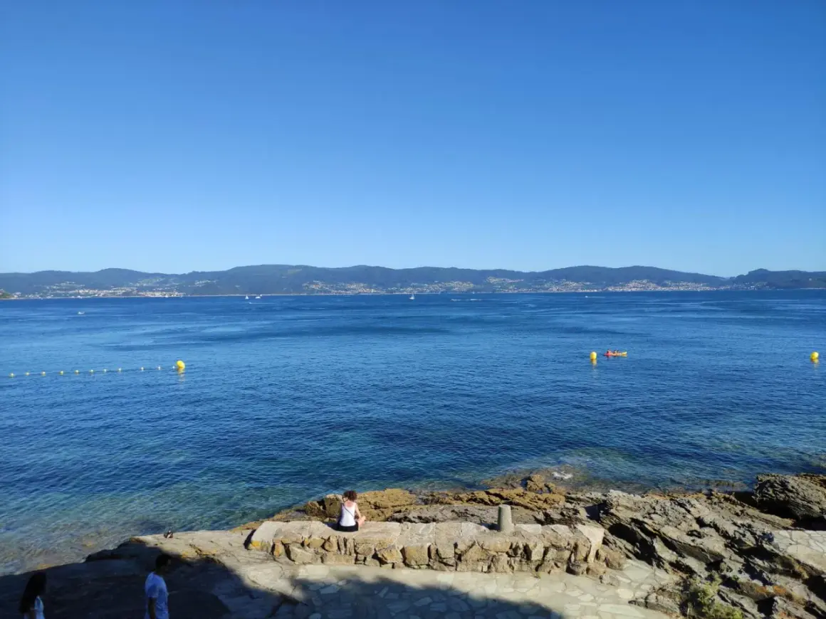 Vista desde punta Faxilda. El mar azul se extiende hasta la costa montañosa, con un kayak solitario y boyas amarillas.
