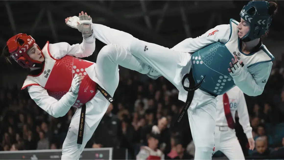Två taekwondo-utövare i strid. Den ena med röd väst och hjälm, den andra med blå väst och hjälm. Båda utför sparkar.