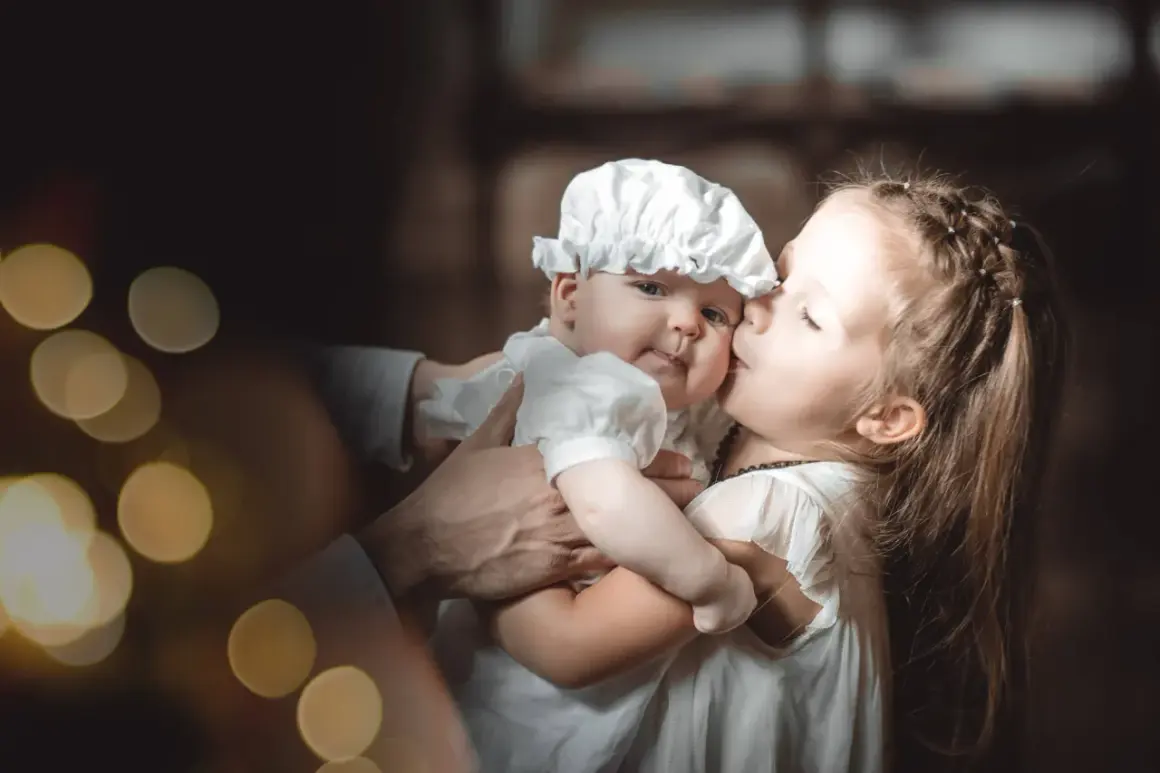 Une sœur embrasse tendrement son bébé, habillé pour baptiser son enfant. Lumières douces et ambiance chaleureuse.