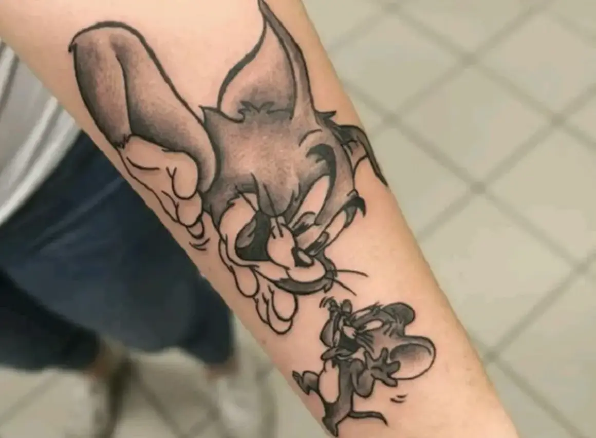 Czarno-biały tatuaż tom i jerry na przedramieniu. Kot Tom próbuje złapać myszkę Jerry, która ucieka.