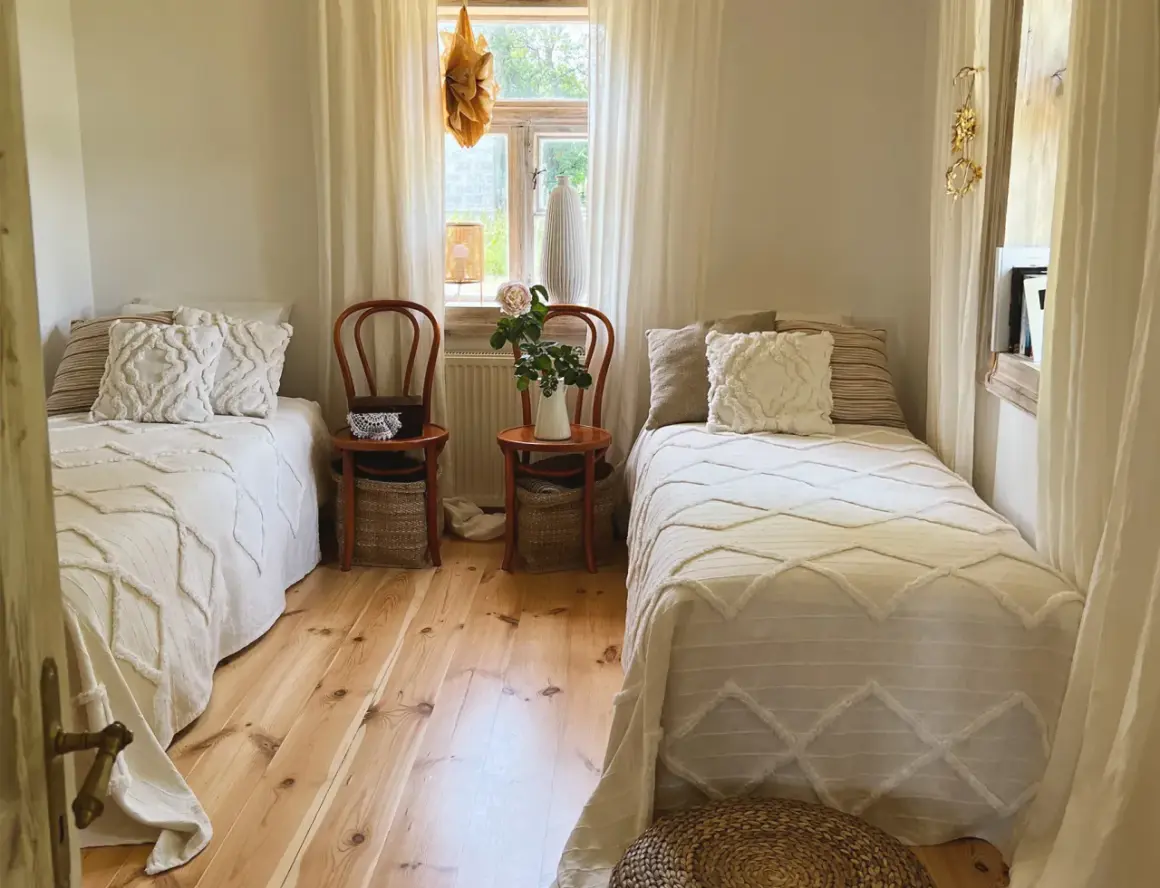 Sypialnia w stylu cottage core z dwoma łóżkami, drewnianą podłogą i oknem z firankami.