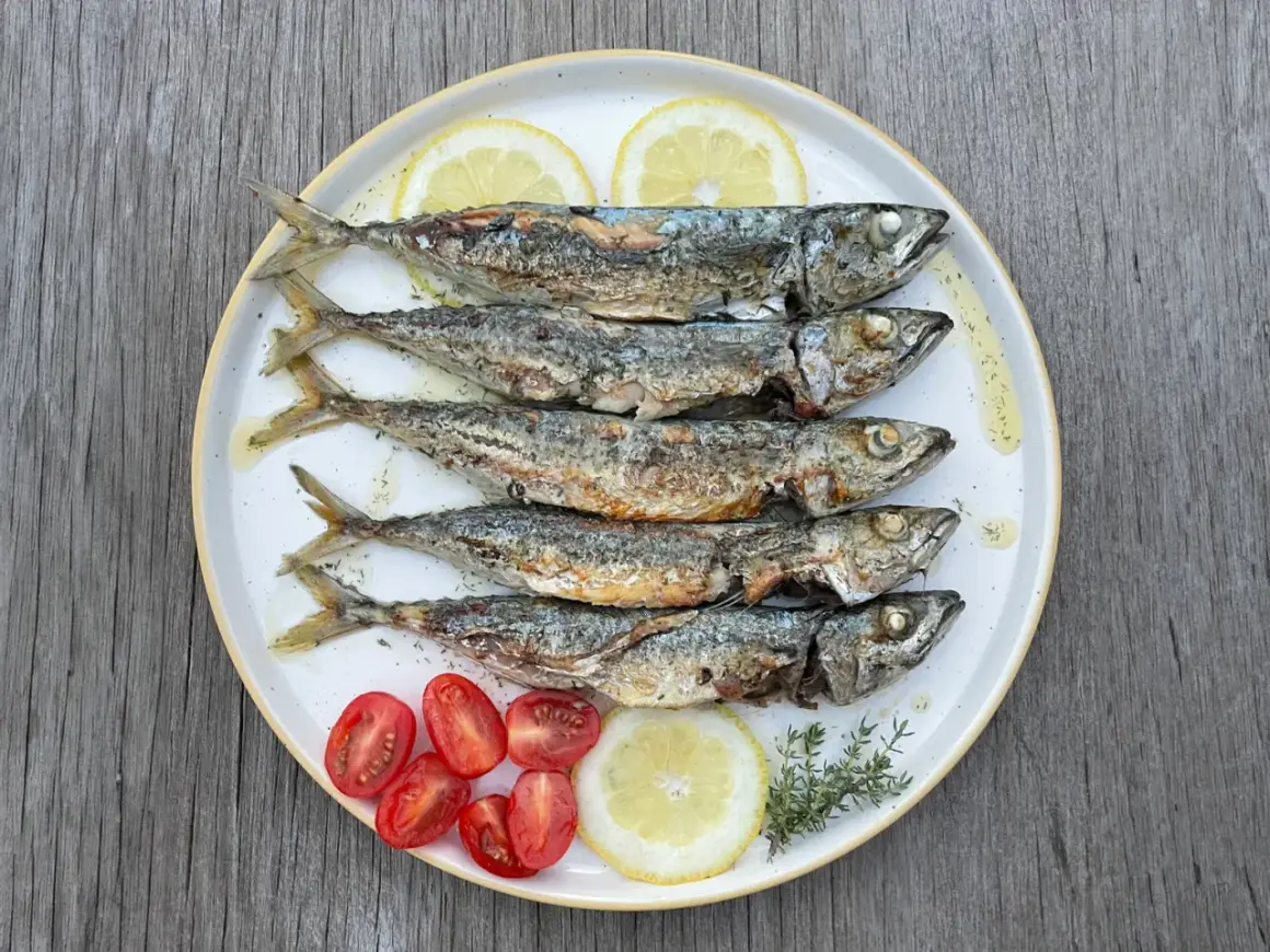 Cinq chinchards poissons grillés, servis sur une assiette avec des quartiers de citron, des tomates cerises et des herbes.