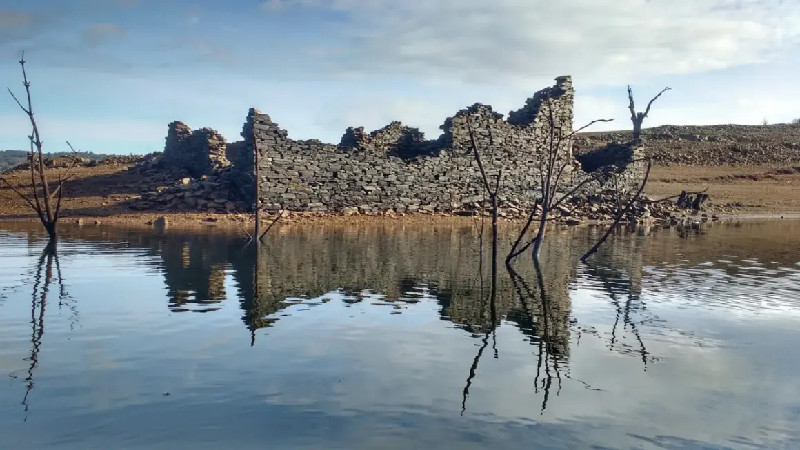 Ruinas de piedra emergen del agua en el embalse de Portodemouros, reflejando su silueta en la calma superficie.