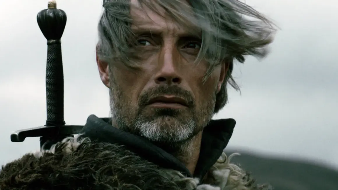Mads Mikkelsen mit Schwert und Pelzmantel, ein Bild, das an seine Rollen in Filmen und Serien erinnert.