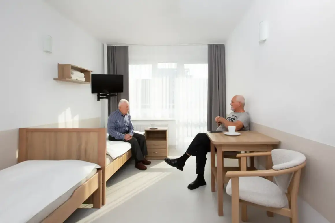 Dwóch starszych panów rozmawia w pokoju sanatorium MSWiA Sopot. Jeden siedzi na łóżku, drugi przy stoliku.