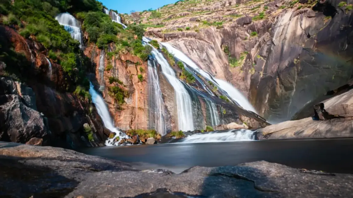 La cascada Fervenza se despliega en múltiples hilos blancos sobre rocas rojizas y verdes, desembocando en un lago sereno.
