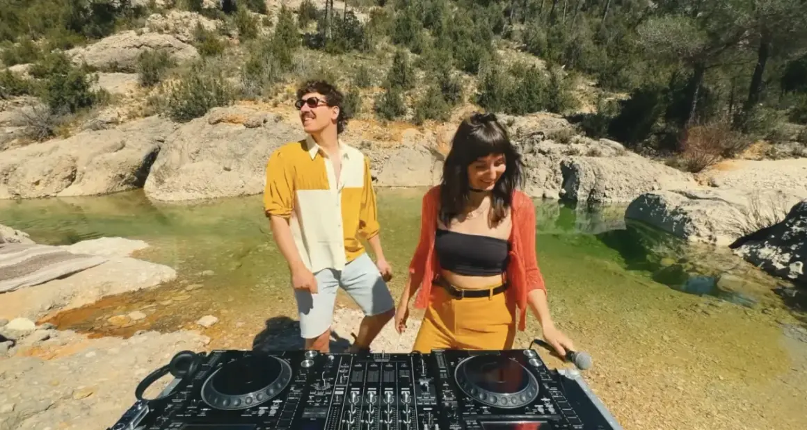 Dwoje DJ-ów przy konsoli w plenerze, nad wodą, w otoczeniu skał i drzew.
