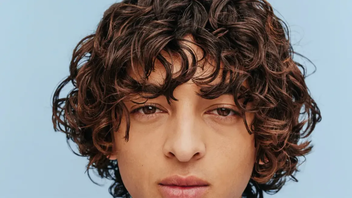 Gros plan sur le visage d'un homme aux cheveux bruns ondulés, type cheveux 2b homme, sur fond bleu clair.