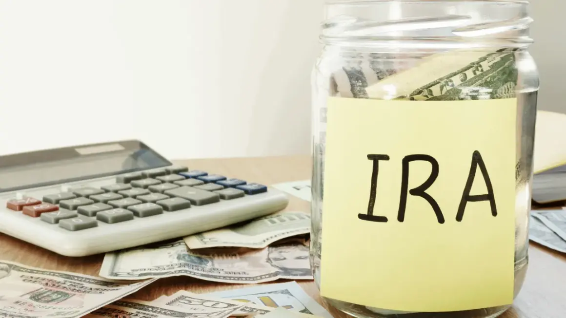 A glass jar labeled "IRA" sits on a table with US dollar bills and a calculator, symbolizing financial planning and the potential for an inherited IRA.