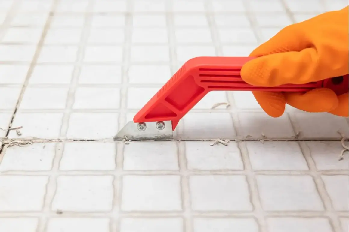 Mano con guanto arancione usa uno strumento rosso per rimuovere lo stucco tra le piastrelle, un passo fondamentale per capire come togliere le piastrelle dal muro.