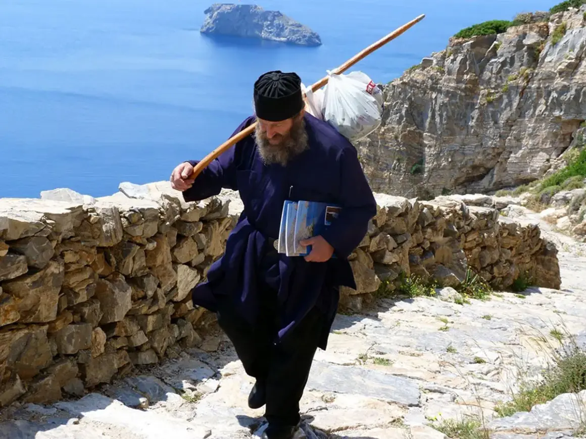 En munk vandrar uppför en stenstig på Amorgos, Grekland, med en stav över axeln och böcker i handen.