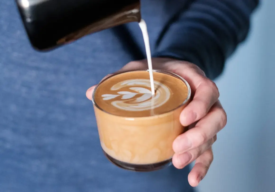 En hand häller mjölk i en kopp för att skapa ett vackert mönster på en flat white.