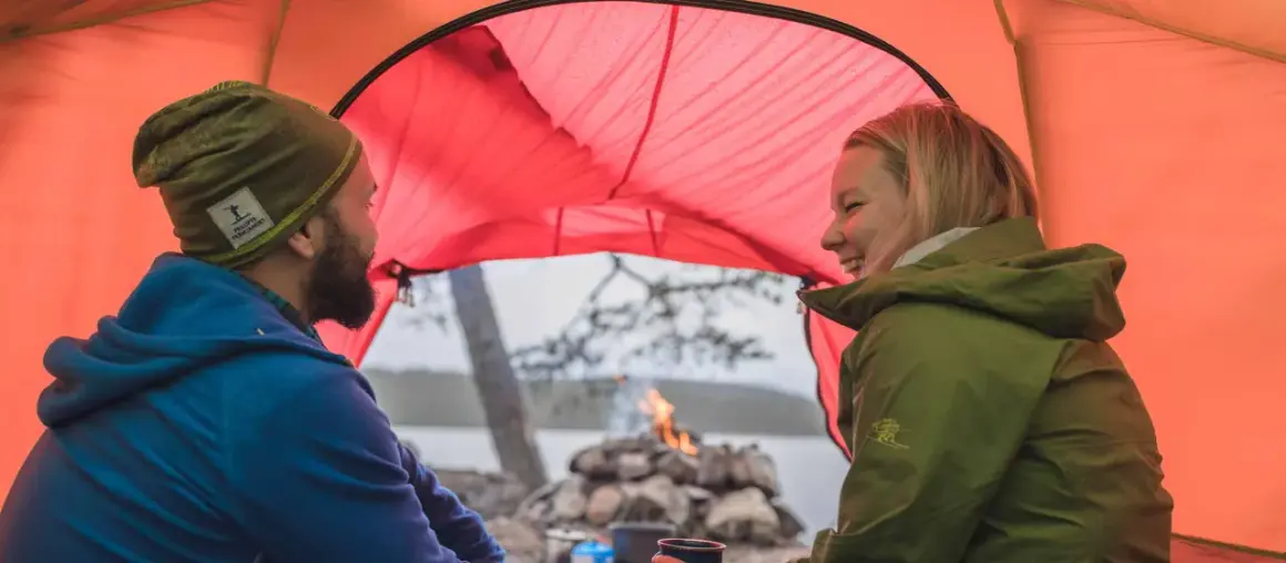 Ett par skrattar inne i ett orange tält. Utanför syns en sjö, träd och en lägereld. Perfekt för att campa i skogen.