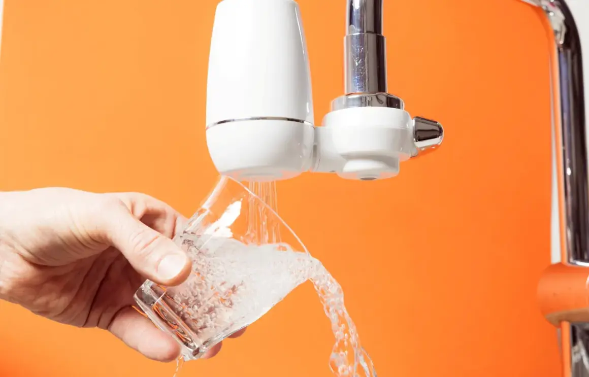 Un robinet avec un filtre blanc verse de l'eau dans un verre. C'est le moment de changer le filtre robinet pour une eau plus pure.