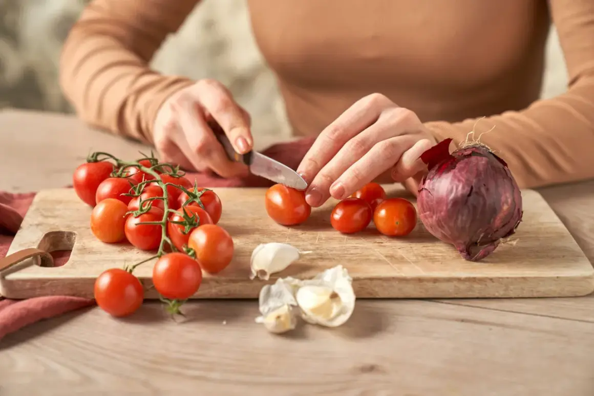 Hände schneiden Tomaten für selbstgemachten Ketchup. Zwiebel und Knoblauch liegen bereit.
