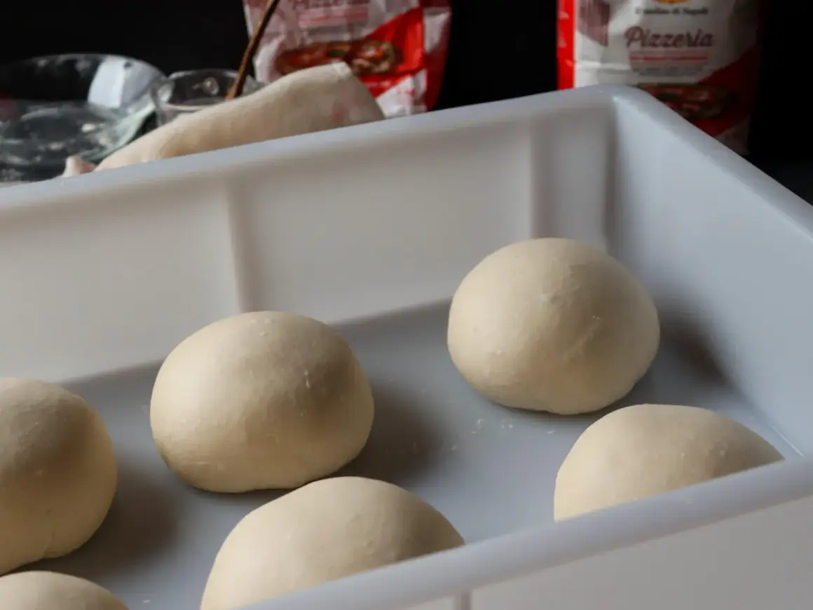 Mehrere Teigkugeln liegen in einer weißen Box, bereit für das Pizzateig einfrieren. Im Hintergrund sind Zutaten und eine Pizzatüte zu sehen.