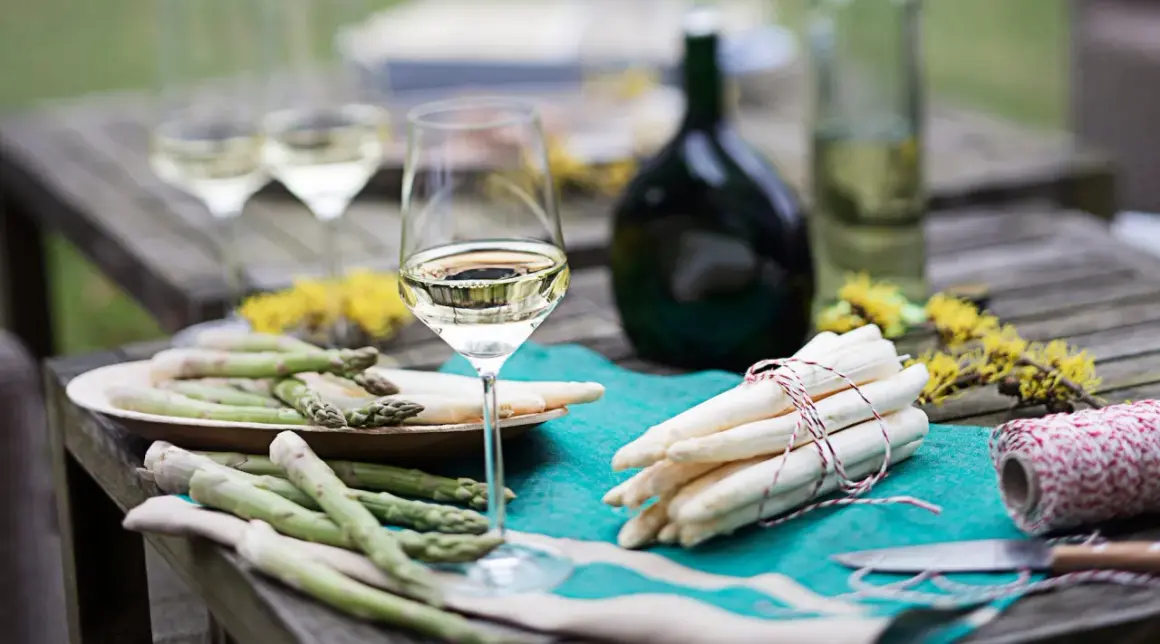 Ein Glas Weißwein steht neben frischem Spargel auf einem Holztisch. Perfekt für den Genuss von Wein zum Spargel.