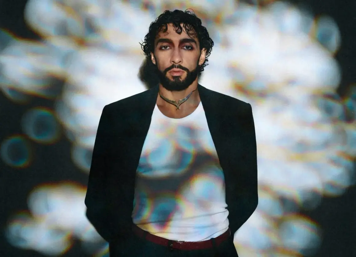 Sofiane Pamart in Berlin, mit dunklem Haar und Bart, trägt einen schwarzen Blazer über einem weißen Shirt. Bunte Lichtreflexe auf seinem Shirt.