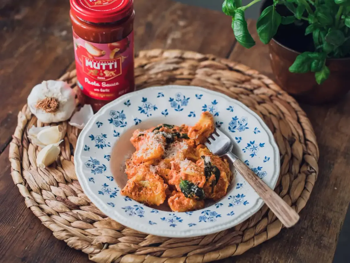En tallrik med krämig tortellini, toppad med parmesan och färska örter. Bredvid står en burk Mutti tomatsås och vitlök, perfekt för ett enkelt tortellini recept.