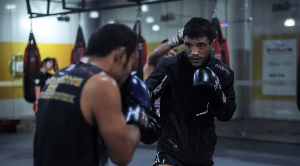 Two men in boxing gloves engage in a spar fight in a gym, with punching bags in the background.