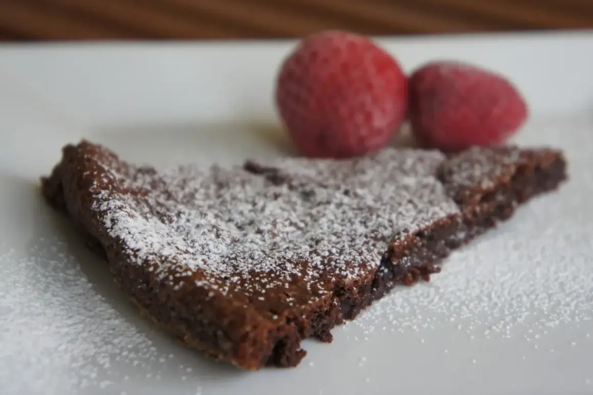 En bit kladdkaka, pudrad med florsocker, med två jordgubbar i bakgrunden. Perfekt för dig som älskar kladdkakecookies utan choklad.