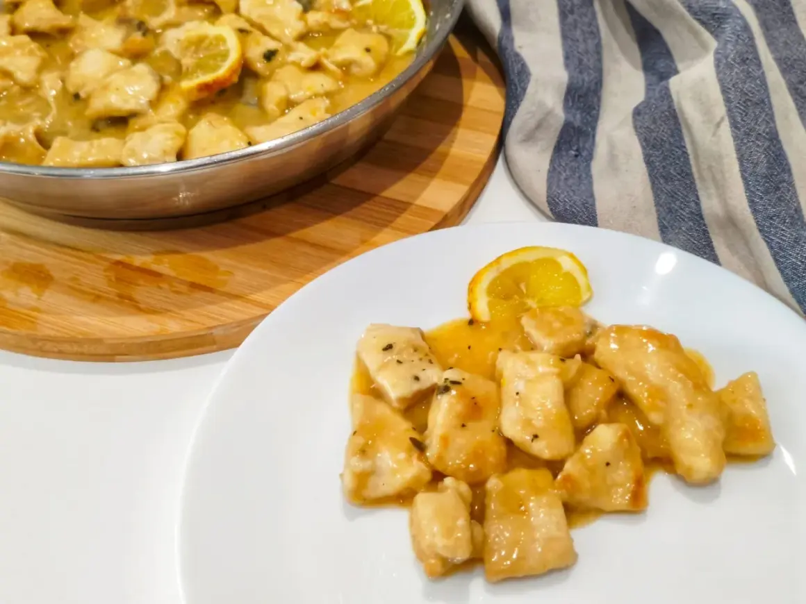 Petto di pollo al limone cremoso servito su un piatto bianco, con una fetta di limone e una padella piena dello stesso piatto sullo sfondo.