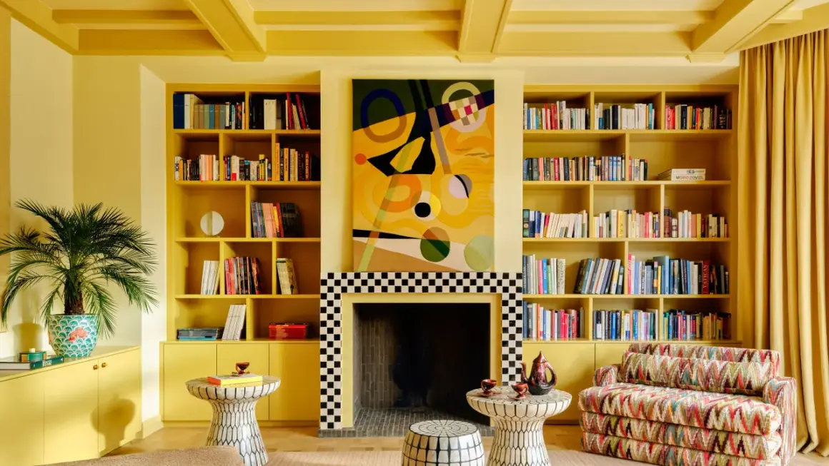 Ein Wohnzimmer in einem gelben Farb-Drenching-Stil mit Bücherregalen, einem Kamin und einem bunten Sofa.