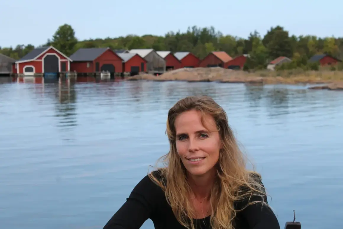 Woman with long blonde hair smiling, with red boathouses on a rocky shore in the background.