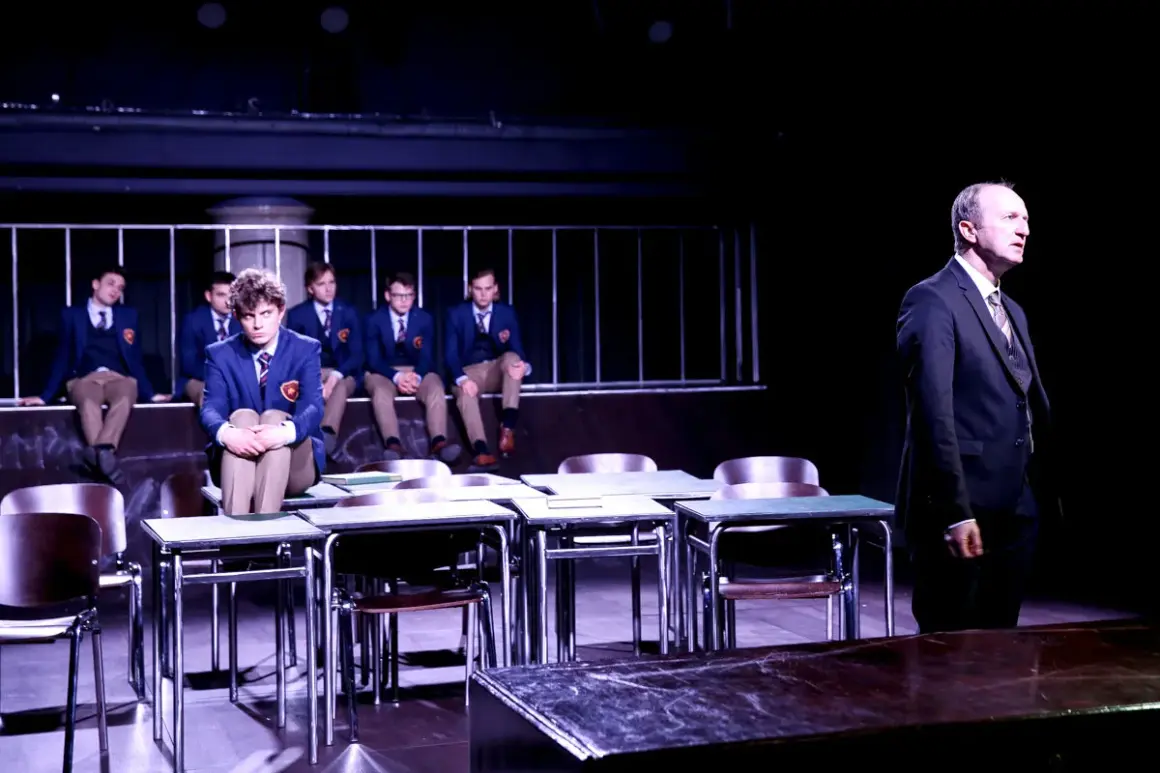 Scena z "Stowarzyszenia Umarłych Poetów" w teatrze. Uczniowie w mundurkach siedzą na ławce, jeden z nich na pierwszym planie. Nauczyciel stoi po prawej.
