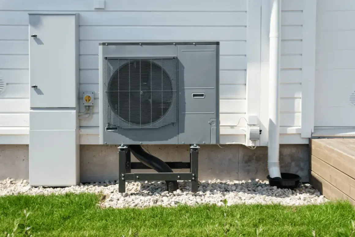 Unità esterna per riscaldamento ad aria, montata su supporti, accanto a un armadio tecnico bianco su una casa con rivestimento in legno.