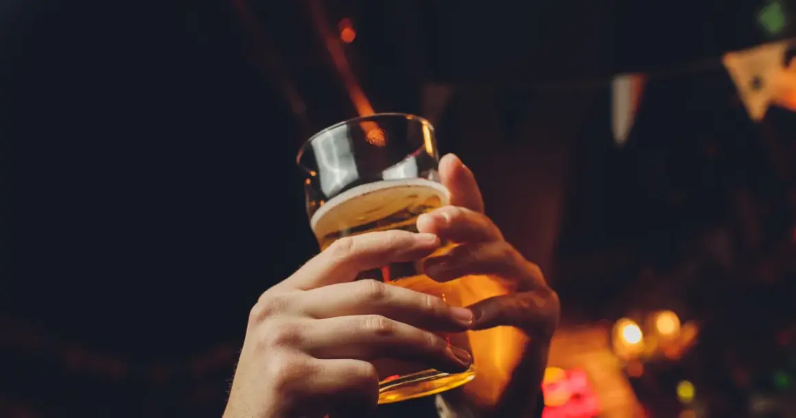 Dwie dłonie trzymające szklankę piwa, w tle rozmyte światła i chorągiewki. Czy piwo tuczy?