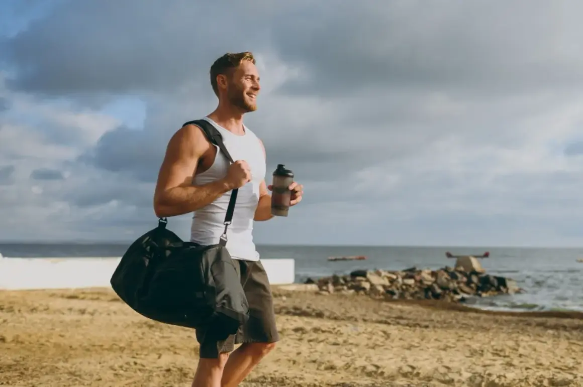 Uśmiechnięty mężczyzna z torbą sportową i bidonem na plaży. Zastanawia się, glutamina co to, czy pomoże mu w regeneracji po treningu.