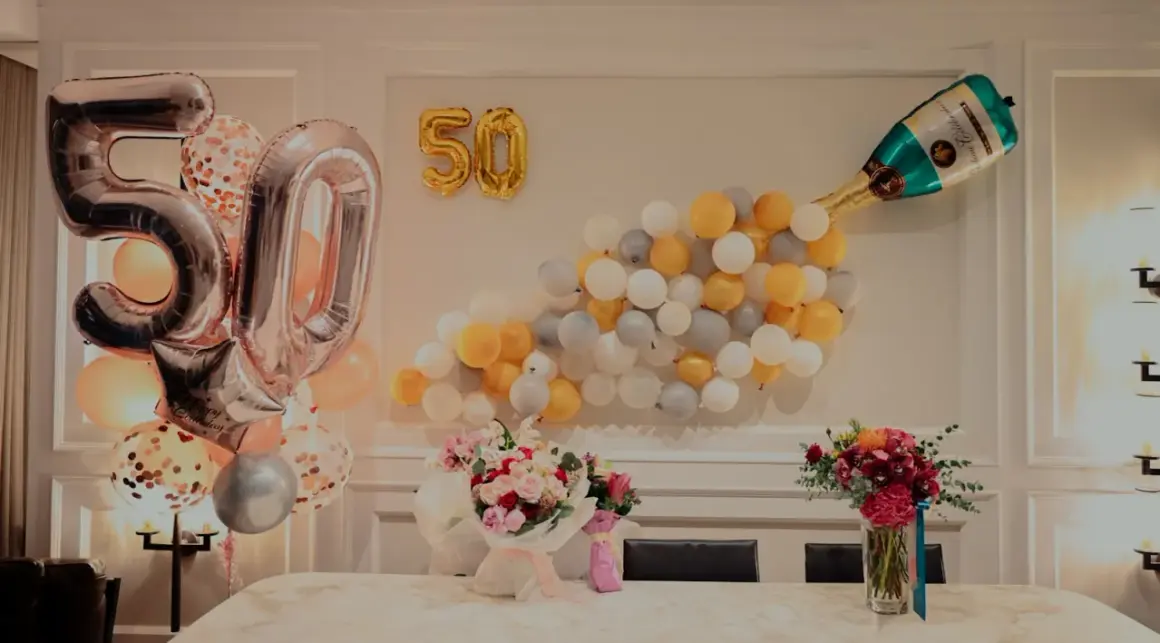 Pięćdziesiąte urodziny żony! Balony w kształcie cyfr 50, szampan i bukiety kwiatów tworzą radosną atmosferę.
