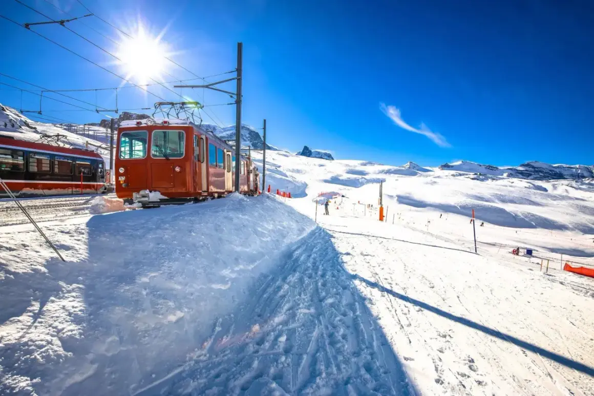 Bernina Express-tåget står parkerat i snön, redo att ta passagerare genom ett snöigt alpint landskap under en klarblå himmel.