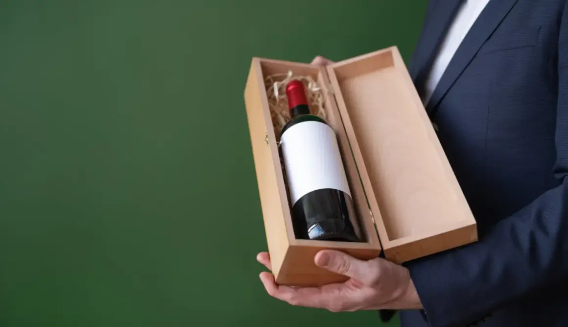 Elegancki mężczyzna w garniturze prezentuje drewniane pudełko z butelką wina wytrawnego czerwonego.