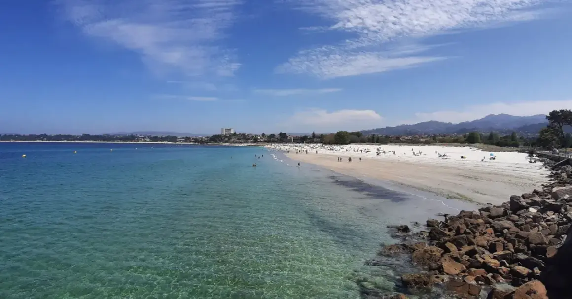 Amplia vista de la playa del Vao en Vigo, con aguas cristalinas, arena blanca y gente disfrutando del sol.